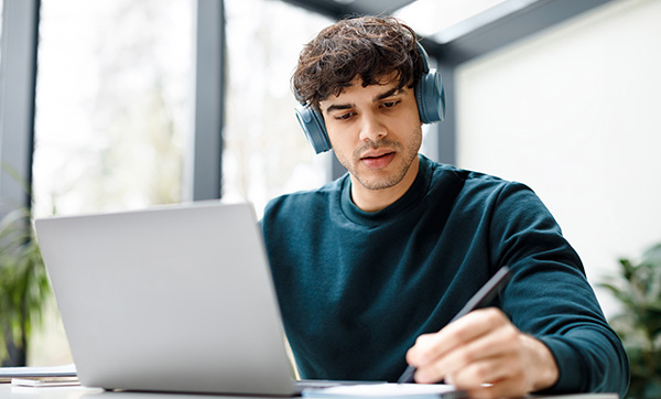 man wearing headphones studying at laptop while taking notes in modern workspace with plants and natural light