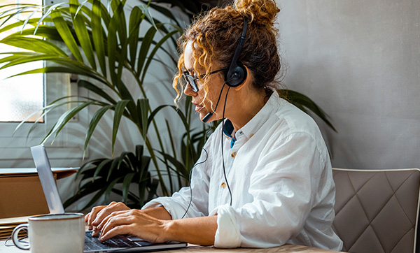 woman working on laptop with headset in home office surrounded by plants engaging in remote communication for virtual meetings