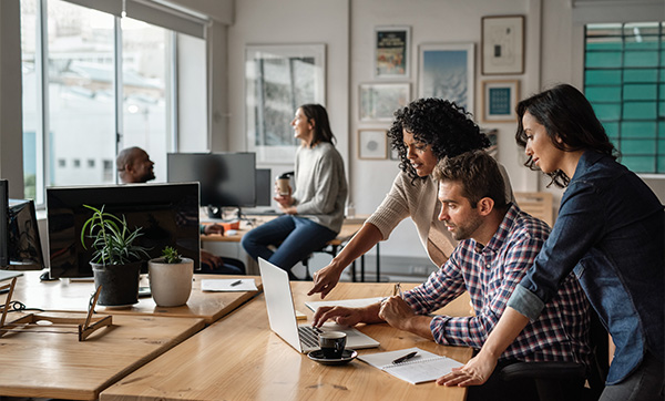 group of three coworkers collaborating in a modern office setting at a laptop with plants and notes on the desk