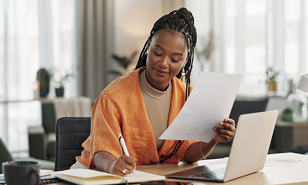 Woman in an orange jacket sitting at a desk reviewing documents and writing notes with a laptop and notebook in the background focused on her work 2 tasks and organization