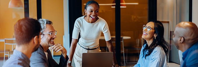 a group of five professionals engaged in a meeting with two women and three men laughing and sharing ideas around a laptop in a modern office setting