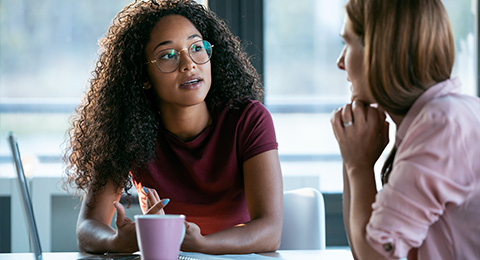 two women engaged in a conversation one woman with curly hair wearing glasses sitting at a table with a notebook and a pink cup discussing ideas in a professional setting three key points being shared