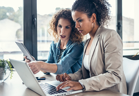 two women collaborating on a laptop discussing ideas with a tablet on a table in a modern workspace